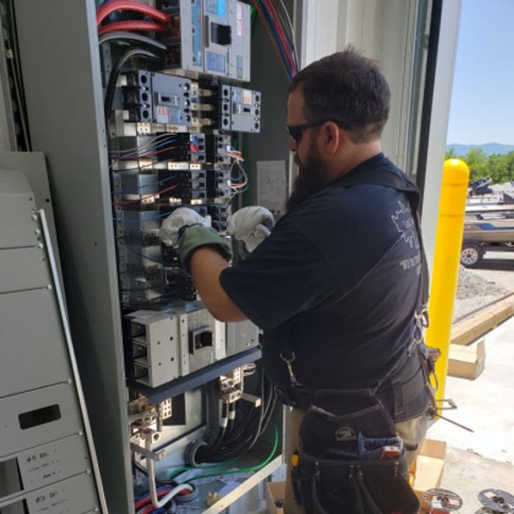 Electrician working on electrical panel with tools and safety gear.