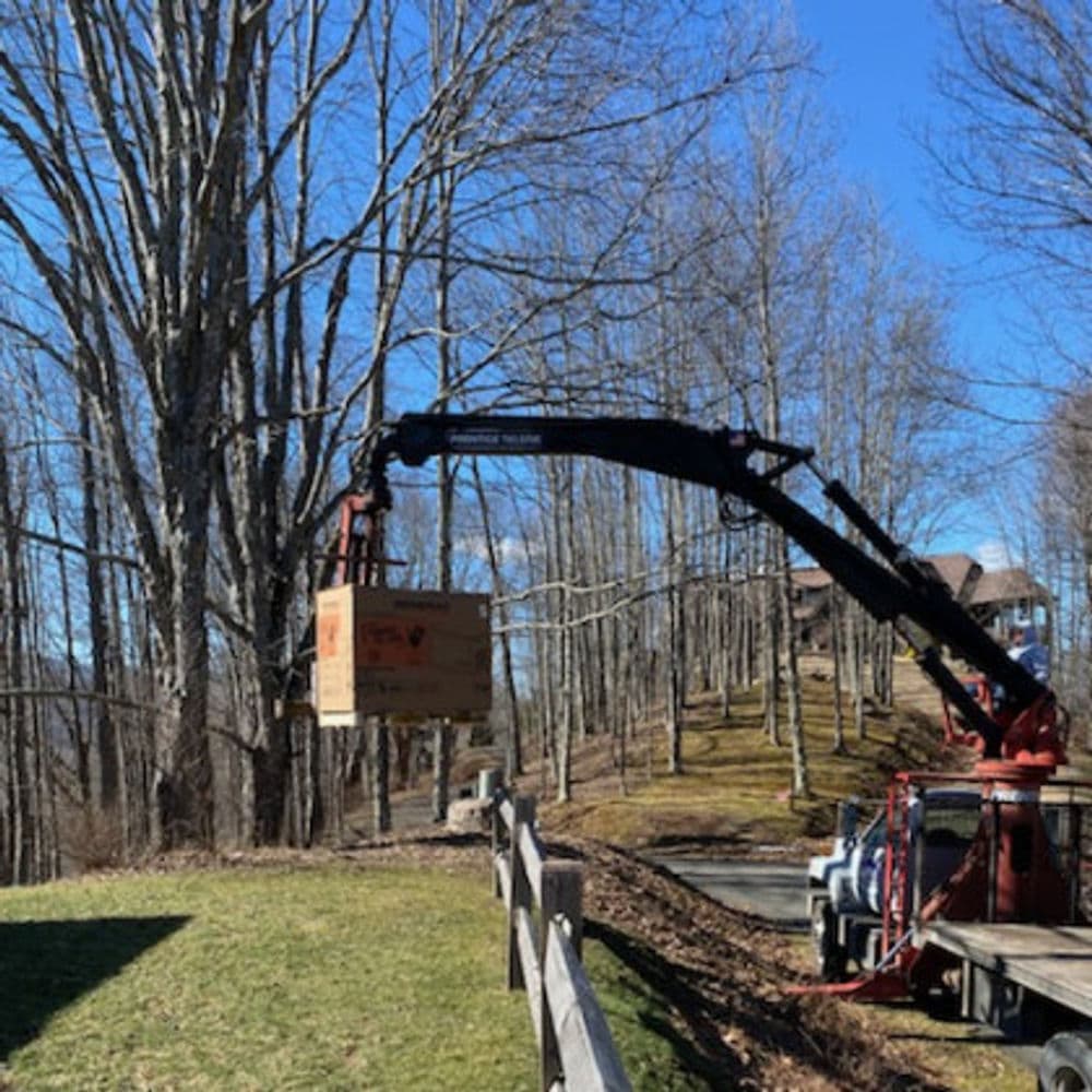 Crane lifting a cardboard box near trees and residential homes on a sunny day.