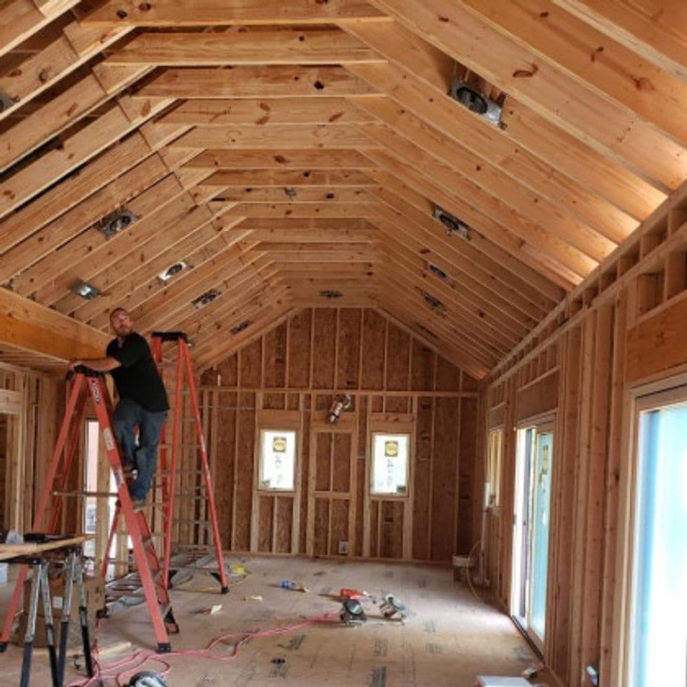 Man on ladder working on interior framing of a house, showcasing wooden beams and open space.